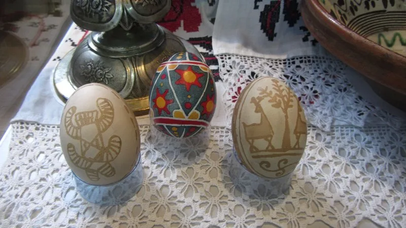 Trois oeufs decores sur un napperon en dentelle : deux graves sur coquille beige avec motifs d'oiseau et de cerf, un peint de couleurs vives